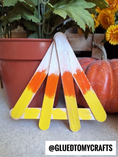 Four craft sticks painted yellow, orange, and white, arranged in a fan shape to resemble craft stick candy corn, are propped up between a potted plant and a pumpkin. The text @GLUEDTOMYCRAFTS is at the bottom of the image.