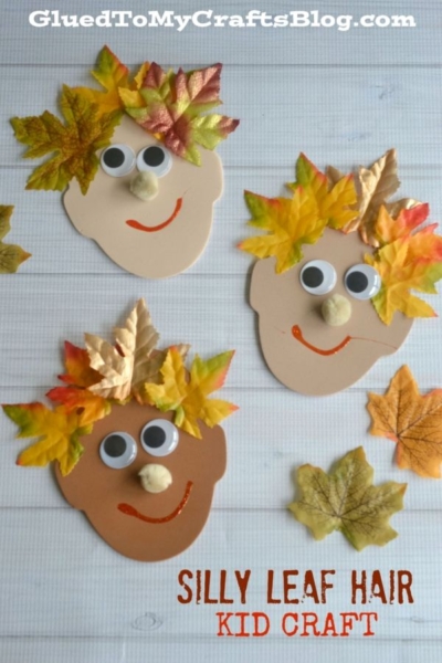 Three paper faces with googly eyes and red smiles, decorated with silly leaf hair in vibrant colors, are arranged on a white surface with extra leaves nearby. The text reads “Silly Leaf Hair Kid Craft.”.