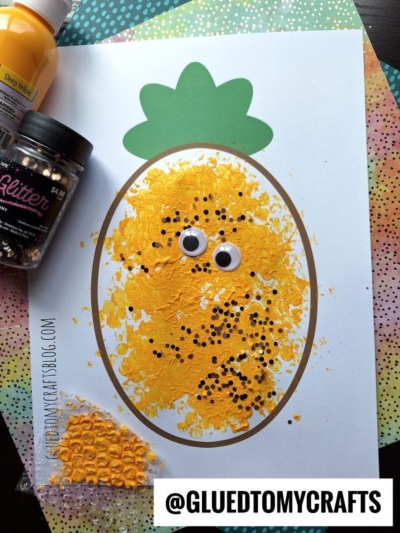 A fun Fruit & Veggie craft, this pineapple project uses yellow paint, googly eyes, and black glitter dots on a printed outline. Craft supplies like yellow paint and glitter are neatly arranged next to the finished artwork.