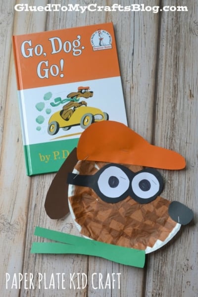 A children’s book titled Go, Dog. Go! by P. D. Eastman sits next to a Go Dog Go Paper Plate craft made from a paper plate, colored paper, and markers, displayed on a wooden surface. Text at the bottom reads Paper Plate Kid Craft.