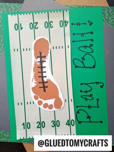 A craft project featuring a brown footprint resembling a football, complete with laces, sits atop a paper football field. For an added personal touch, incorporate a handprint football goal keepsake to frame your creation. The words Play Ball! are written in black to the right, while @GLUEDTOMYCRAFTS is displayed at the bottom.