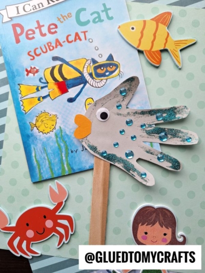 A Pete the Cat Scuba-Cat book is surrounded by ocean-themed cutouts, including a crab, mermaid, and colorful handprint fish puppets—one made from a glittery handprint on a wooden stick. @GLUEDTOMYCRAFTS is at the bottom.