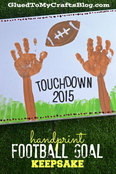 A craft project featuring two brown handprints as a handprint football goal keepsake, a painted football, and the text “TOUCHDOWN 2015,” set on a grassy background with “handprint FOOTBALL GOAL KEEPSAKE” included.