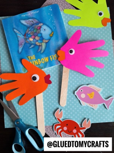A children’s book titled The Rainbow Fish sits on patterned paper surrounded by colorful handprint fish puppets made from hand-shaped paper, googly eyes, and craft sticks. Scissors and cartoon fish and crab cutouts are also nearby.
