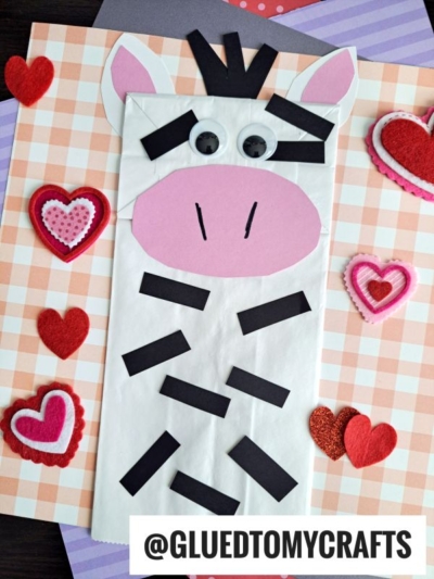 A lunch sack zebra craft with a pink oval snout, googly eyes, and black paper stripes sits on a checkered background decorated with felt hearts. @GLUEDTOMYCRAFTS is printed at the bottom.