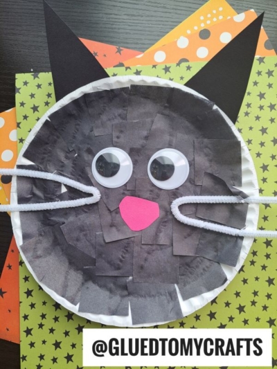 A round paper black cat face craft made from paper, pipe cleaners for whiskers, big googly eyes, and a pink nose, displayed on star-patterned paper. Text reads @GLUEDTOMYCRAFTS.