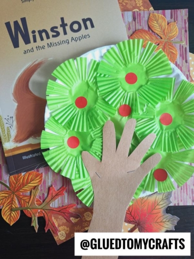 A children’s book, paper hand cutout, and a colorful cupcake liner apple tree craft, featuring green cupcake liner apples on the branches with paper leaves scattered around.
