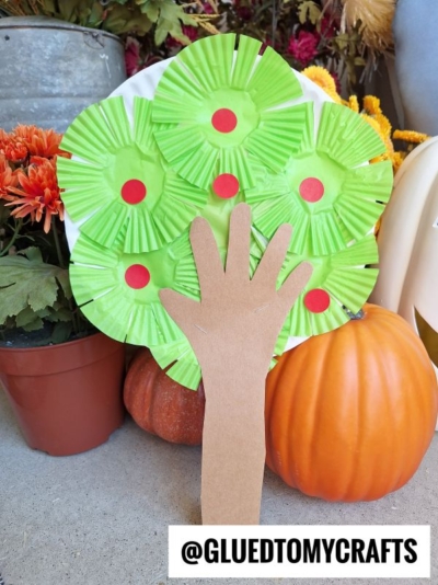 A handprint-shaped brown paper forms a tree trunk, topped with green cupcake liners and red dots to create a charming cupcake liner apple tree. The craft is placed among fall pumpkins and flowers. Text reads @GLUEDTOMYCRAFTS.