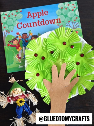 A children’s book titled Apple Countdown, a cupcake liner apple tree craft made with green liners and red dots, and a small scarecrow doll are arranged on a dark table. @GLUEDTOMYCRAFTS is written at the bottom.
