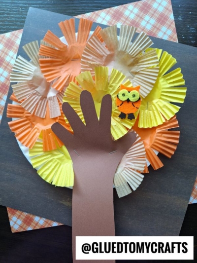 A fall tree craft made from brown paper shaped like a hand for the trunk, colorful cupcake liners for leaves reminiscent of a cupcake liner apple tree, and a small orange owl decoration, all on a dark background.