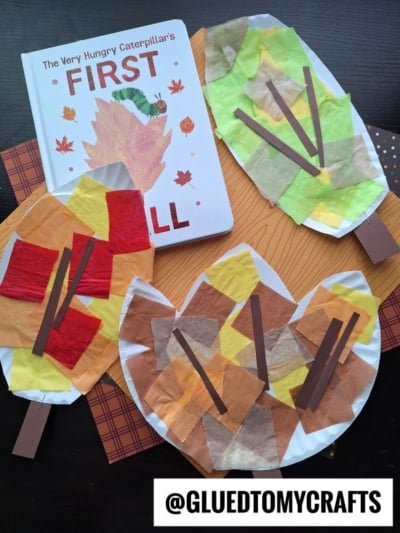 A children’s book, The Very Hungry Caterpillar’s First Fall, is surrounded by three paper plate leaf crafts decorated with colored tissue paper and brown paper strips. The handle @GLUEDTOMYCRAFTS is at the bottom.