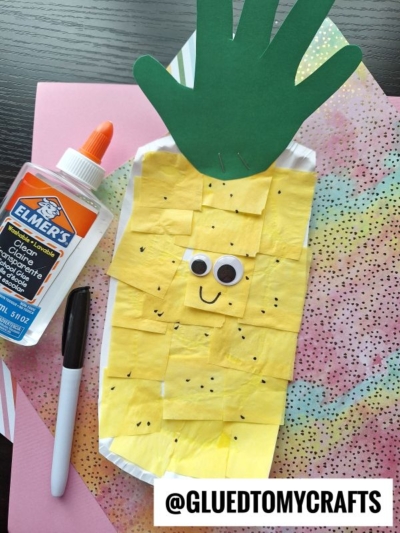 A pineapple Fruit & Veggie craft made from yellow paper squares and a green handprint on a paper plate, with googly eyes and a smile, sits on colorful paper next to glue and a black marker. @GLUEDTOMYCRAFTS is written at the bottom.