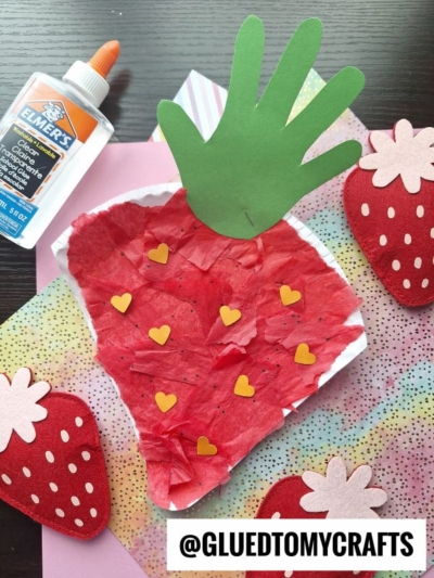 A strawberry craft made from red tissue paper, yellow heart shapes, and a green paper hand for the leaf sits on colorful paper—perfect for Fruit & Veggie crafts. Elmer’s glue and felt strawberries are nearby. Text reads @GLUEDTOMYCRAFTS.