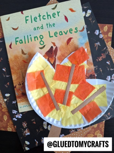 A children’s book titled “Fletcher and the Falling Leaves” lies on autumn-themed paper, topped with a paper plate leaf craft decorated in orange, yellow, and brown tissue paper and craft sticks. @GLUEDTOMYCRAFTS is written at the bottom.