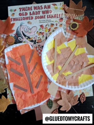 Children’s fall craft scene featuring a paper plate leaf and a paper leaf, accented with orange and yellow construction paper strips, felt leaves, and the book “There Was An Old Lady Who Swallowed Some Leaves!” in the background. @GLUEDTOMYCRAFTS tag at bottom.