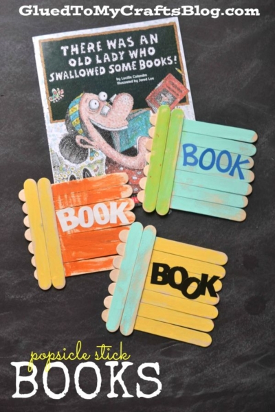 Popsicle stick books shaped like tiny novels, painted in orange, yellow, and blue with BOOK on each. Behind them is the book There Was an Old Lady Who Swallowed Some Books! resting on a dark surface.