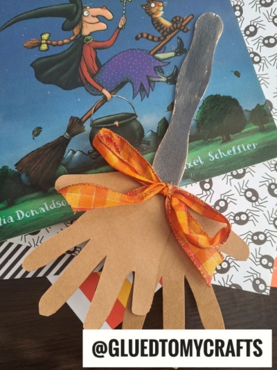A handmade popsicle stick broom craft, created from brown paper hand cutouts and tied with an orange ribbon, sits atop the book “Room on the Broom.” A tag reads @GLUEDTOMYCRAFTS.