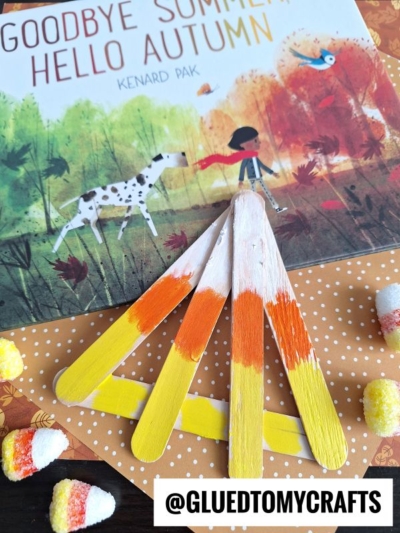 A children’s book titled “Goodbye Summer, Hello Autumn” rests on a patterned surface beside a popsicle stick candy corn craft, surrounded by candy corn pieces and a tag reading @GLUEDTOMYCRAFTS.