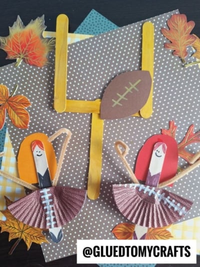 Handmade cheerleader dolls with smiling faces, made from clothespins and cupcake liners, stand in front of a popsicle stick mini football goal and football, surrounded by autumn leaves and polka-dot paper.