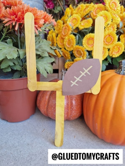 A popsicle stick mini football goal made from yellow sticks, with a brown paper football, stands among pumpkins and potted orange and yellow flowers. Text reads @GLUEDTOMYCRAFTS.