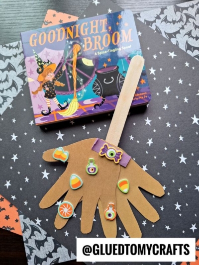 A children’s book titled Goodnight, Broom sits next to a handprint paper craft broomstick—complete with a popsicle stick broom handle—decorated with Halloween stickers on a dark, starry background, reminiscent of the magical adventures in Room on the Broom. @GLUETOMYCRAFTS is written at the bottom.