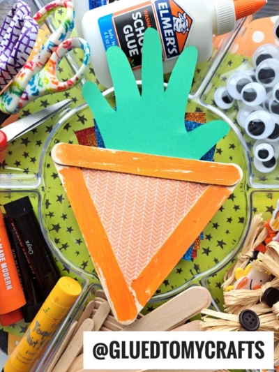 A handmade carrot craft, perfect for Fruit & Veggie crafts, made with orange paper, a green handprint top, and craft sticks sits on a tray surrounded by glue, scissors, googly eyes, and glue sticks. Text: @GLUEDTOMYCRAFTS.