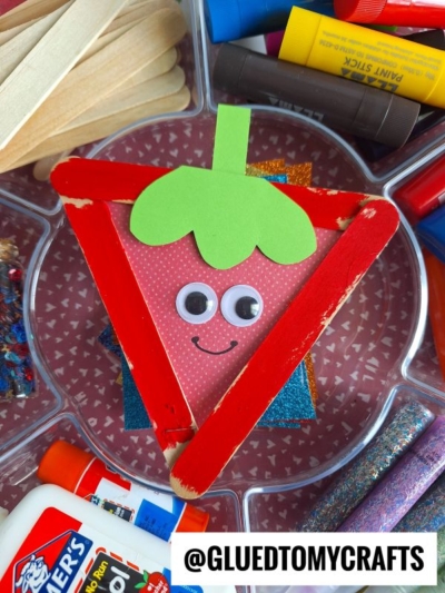 A strawberry craft made from red craft sticks, green paper, googly eyes, and a smiling face, surrounded by glue, glitter, markers, and popsicle sticks. Perfect for Fruit & Veggie crafts! @GLUEDTOMYCRAFTS is labeled at the bottom.