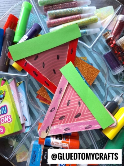Two triangular watermelon Fruit & Veggie crafts made from popsicle sticks and paper are surrounded by colorful craft supplies like glue sticks, glitter, and markers. A label at the bottom reads @GLUEDTOMYCRAFTS.
