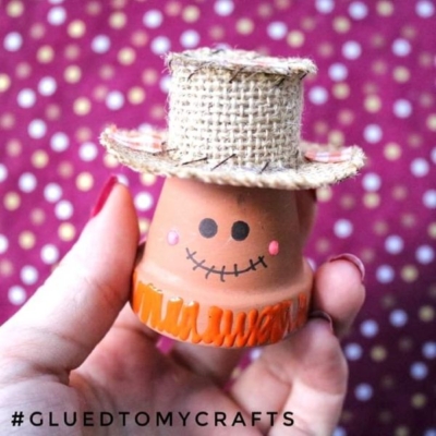 A hand holds a small, painted terra cotta pot scarecrow, decorated as a smiling figure with a burlap hat. A dotted background is visible. #GLUEDTOMYCRAFTS.