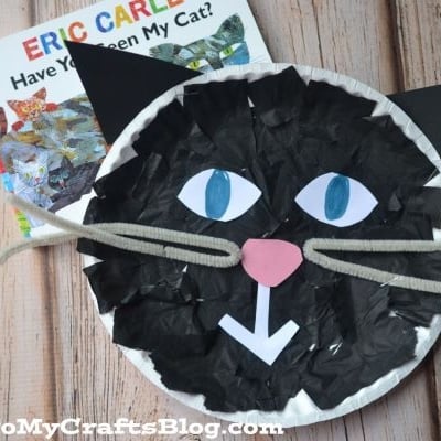 A children’s book titled Have You Seen My Cat? by Eric Carle sits next to a paper black cat craft made from a paper plate, tissue paper, and yarn whiskers on a wooden surface.