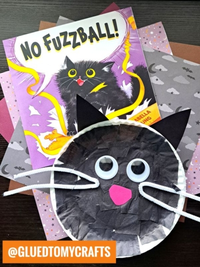 A children’s book titled No Fuzzball! with a black cat on the cover sits behind a paper black cat face craft made from a paper plate, black and pink paper, googly eyes, and white pipe cleaners. Patterned papers are underneath.