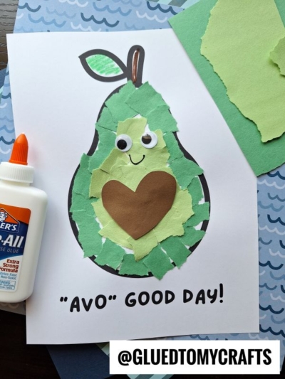 A fun Fruit & Veggie craft: a torn paper avocado with googly eyes, a smile, and a brown heart pit on white paper. A glue bottle sits nearby. Text below reads “‘AVO’ GOOD DAY!” and “@GLUEDTOMYCRAFTS.”.