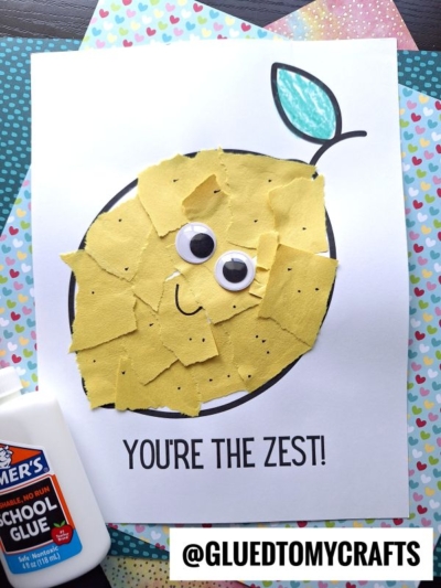 A cute Fruit & Veggie craft made with torn yellow paper pieces, googly eyes, and a drawn stem and leaf on white paper. Text below reads “YOU’RE THE ZEST!” A bottle of glue sits nearby, with colorful paper in the background.