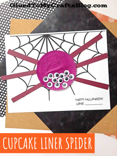 A fun Halloween craft featuring a cupcake liner spider with black strip legs and googly eyes, set on paper with a hand-drawn web and “Happy Halloween” message.