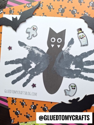 A Halloween craft featuring a black paper owl with wings made from painted handprints, surrounded by cartoon ghosts, bats, stars, and a special handprint bat keepsake set on festive orange and purple backgrounds.