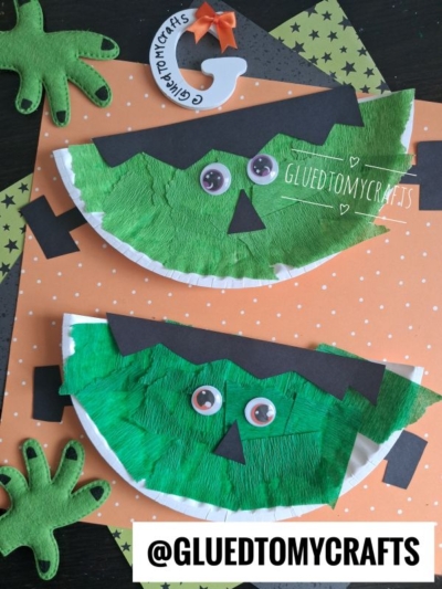 Two paper plate frankenstein crafts painted green with black shapes and googly eyes, resembling Frankenstein faces. Placed on orange polka-dot paper with felt Frankenstein hands and a small paper pumpkin decoration. @GLUEDTOMYCRAFTS is written at the bottom.