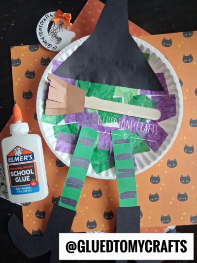 A paper plate melted witch craft with green and purple paper strips, black hat, broom, and dangling legs on a Halloween-themed background. Elmer’s glue and a decorated foam cup are nearby. @GLUEDTOMYCRAFTS is at the bottom—perfect for creating your own whimsical paper plate melted witch this spooky season!.