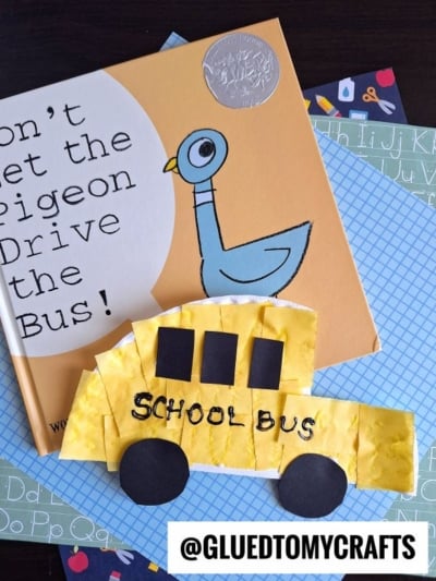 A children’s book titled Don’t Let the Pigeon Drive the Bus! is next to a cheerful paper plate school bus craft, all placed on blue patterned sheets. @GLUETOMYCRAFTS is at the bottom.