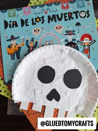 A Día de los Muertos book with a creative paper plate skull craft made from white tissue paper and black paper, featuring eyes and a nose, displayed next to a tag reading @GLUEDTOMYCRAFTS.