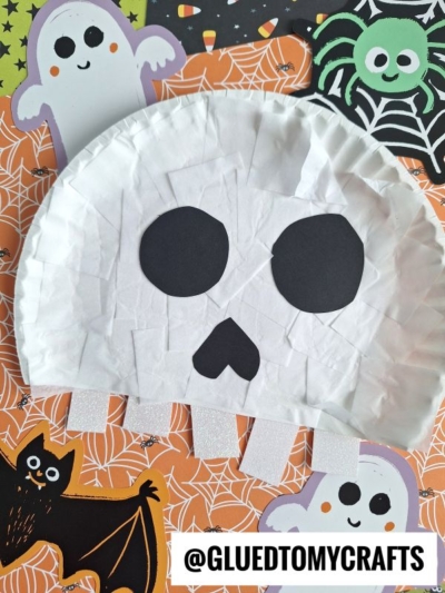 A paper plate skull craft with black paper eyes and nose sits on a Halloween-themed background featuring ghosts, a spider, and a bat. This spooky yet fun paper skull project is perfect for holiday crafting, with the text @GLUEDTOMYCRAFTS appearing at the bottom.