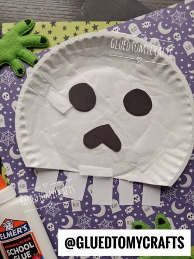 A paper plate ghost craft with black eyes and mouth on star-patterned paper, felt green hands, glue, Halloween-themed cards, and a playful twist inspired by a classic paper plate skull. Text reads @GLUEDTOMYCRAFTS.