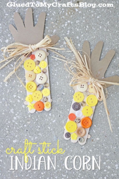 Two hand-shaped paper cutouts are attached to popsicle sticks decorated with yellow, white, and orange buttons to resemble craft stick corn, with raffia tied around the top as husks, on a speckled surface.