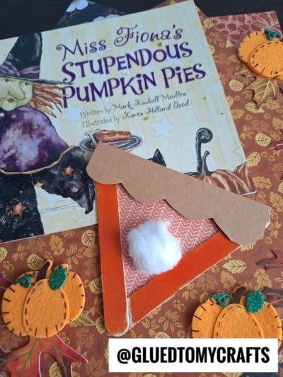 A craft stick pumpkin pie slice with an orange body, tan paper crust, and cotton ball dollop, resting on a copy of Miss Fiona’s Stupendous Pumpkin Pies surrounded by felt pumpkin decorations.