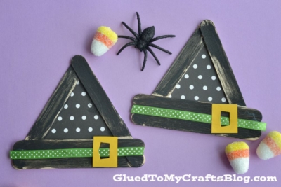 Two popsicle stick witch hats made from black popsicle sticks are adorned with green polka-dot ribbon and yellow buckles, set against a purple background. Candy corn pieces and a black spider are arranged nearby.