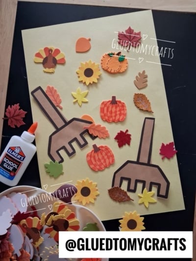 A fall-themed raking leaves kid craft with paper rakes, pumpkins, leaves, and sunflowers on yellow paper. Elmers glue and autumn-shaped foam stickers are nearby. Username @GLUETOMYCRAFTS appears on the image.