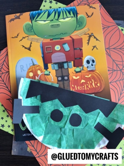 A children’s Halloween book with a cartoon Frankenstein on the cover, surrounded by pumpkins and bats, sits next to a playful paper plate Frankenstein craft made from a paper plate. Orange paper with spiderweb designs is underneath.