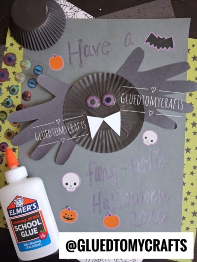 A Halloween craft featuring a cupcake liner bat made from black cupcake liners and handprint cutouts on gray paper. Surrounding decorations include pumpkins, skulls, and glue. Text reads, Have a fang-tastic Halloween 2026.