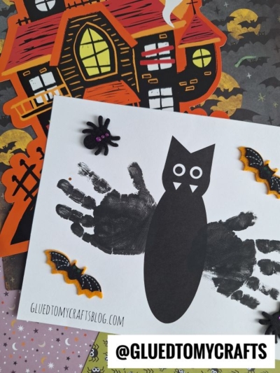 A Halloween craft project featuring a paper owl made from black handprints, with paper wings, eyes, and a beak. The artwork is surrounded by Halloween-themed decorations like a haunted house, bats, and a handprint bat keepsake.