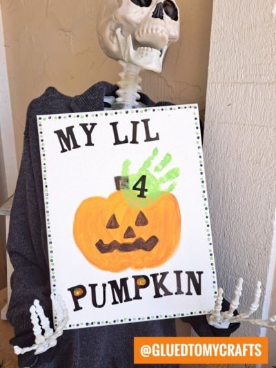 A skeleton holds a sign reading MY LIL 4 PUMPKIN with a painted handprint pumpkin keepsake, featuring a green handprint stem and jack-o-lantern face. The sign is bordered with small colorful dots.