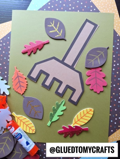 A raking leaves kid craft featuring a paper rake, colorful paper leaves, and a glue stick is centered on a green sheet. The Instagram handle @GLUEDTOMYCRAFTS appears at the bottom.